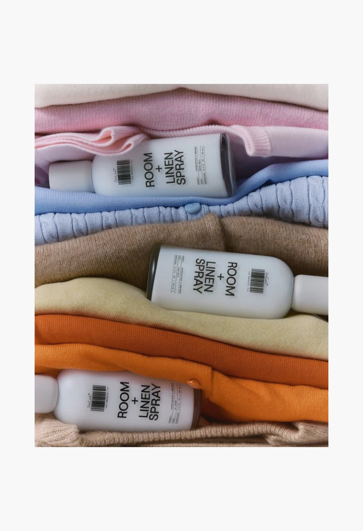 GLAM MODA Xtra Milk room and linen spray bottles resting on a stack of folded pastel and warm-toned knit sweaters.